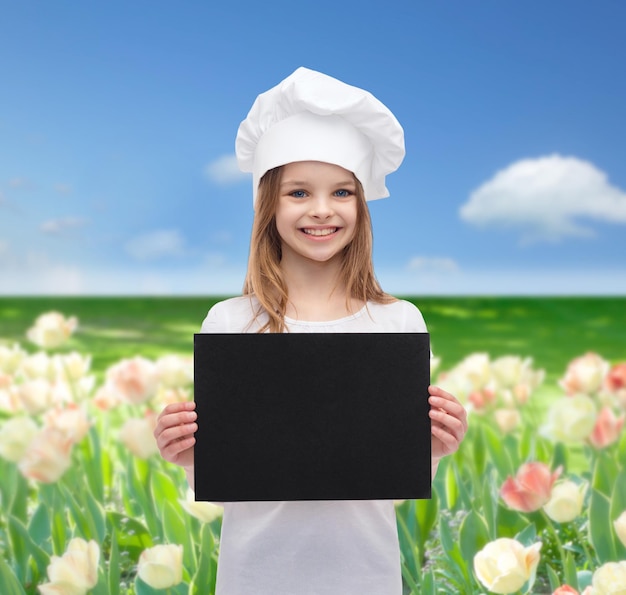 Kochen, Kindheit, Werbung und Personenkonzept - lächelndes kleines Kochmädchen, Köchin oder Bäcker mit leerem schwarzem Papier über blauem Himmel und Blumenfeldhintergrund