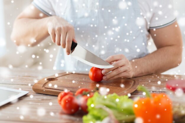 Kochen, Menschen, vegetarisches Essen und Wohnkonzept - Nahaufnahme eines Mannes, der mit einem großen Messer Tomaten und anderes Gemüse auf einem Schneidebrett hackt