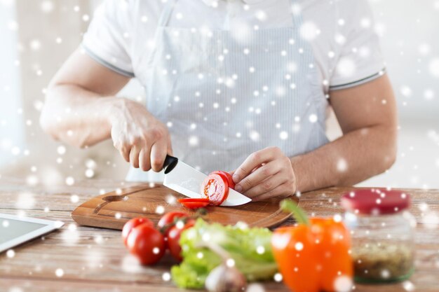 Kochen, Menschen, vegetarisches Essen und Wohnkonzept - Nahaufnahme eines Mannes, der mit einem großen Messer Tomaten und anderes Gemüse auf einem Schneidebrett hackt