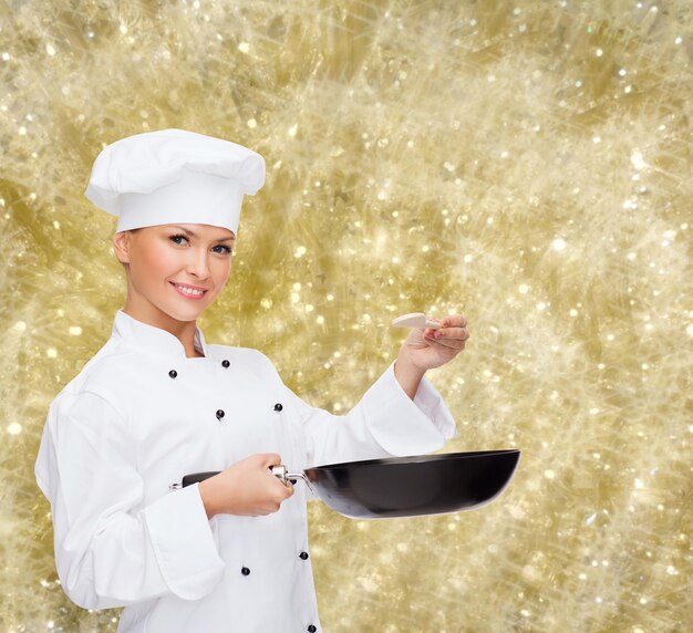 Kochen, Urlaub, Menschen und Food-Konzept - lächelnde Köchin mit Pfanne und Löffel, die Essen auf gelbem Hintergrund schmeckt