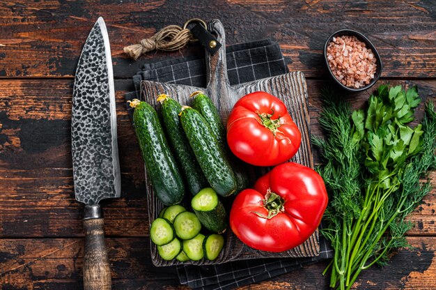 Kochen von grünem Gemüsesalat mit Tomaten, Gurken, Petersilie, Kräutern. Dunkler hölzerner Hintergrund. Ansicht von oben.