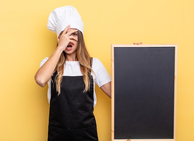 Kochfrau, die schockiert, verängstigt oder verängstigt aussieht und das Gesicht mit dem Handtafel-Menükonzept bedeckt