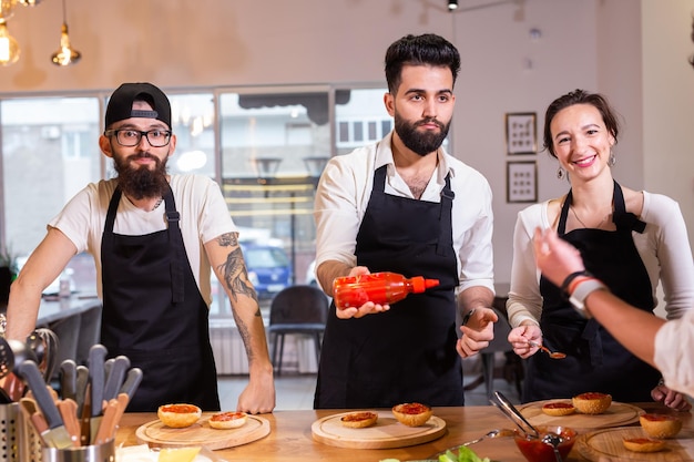 Kochkurs: Ein leitender männlicher Koch in Kochuniform bringt jungen Leuten im Kochkurs bei, Zutaten für Gerichte in der Restaurantküche zuzubereiten und zu mischen