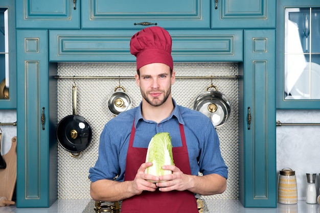 Kochmann hält Chinakohl in der Küche. Koch mit rotem Hut, Schürze mit Gemüse. Vegetarisch, Gesundheit, Ernährung, Vitamin. Essen, Kochen, Küche, Menü, Gerichte, Rezepte.
