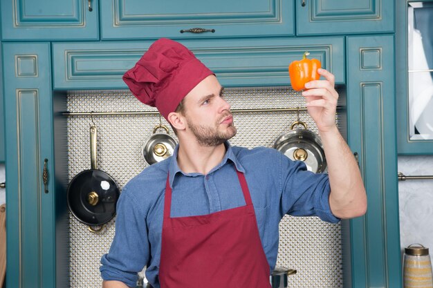 Kochmann hält Paprika in der Küche. Koch mit rotem Hut, Schürze mit Gemüse. Vegetarisch, Gesundheit, Ernährung, Vitamin. Essen, Kochen, Küche, Menü, Gerichte, Rezepte.