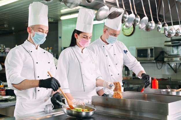 Köche in Schutzmasken und Handschuhen bereiten Speisen in der Küche eines Restaurants oder Hotels zu.