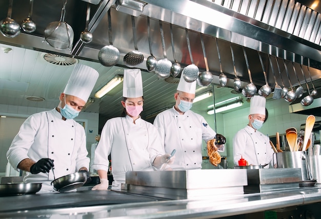 Köche in Schutzmasken und Handschuhen bereiten Speisen in der Küche eines Restaurants oder Hotels zu.