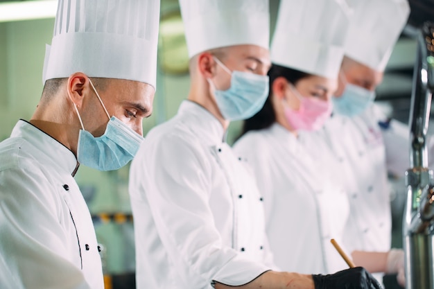 Köche in schutzmasken und handschuhen bereiten speisen in der küche eines restaurants oder hotels zu.