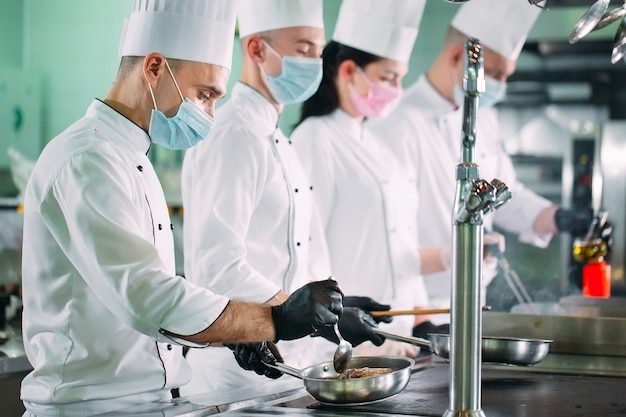 Köche in Schutzmasken und Handschuhen bereiten Speisen in der Küche eines Restaurants oder Hotels zu.