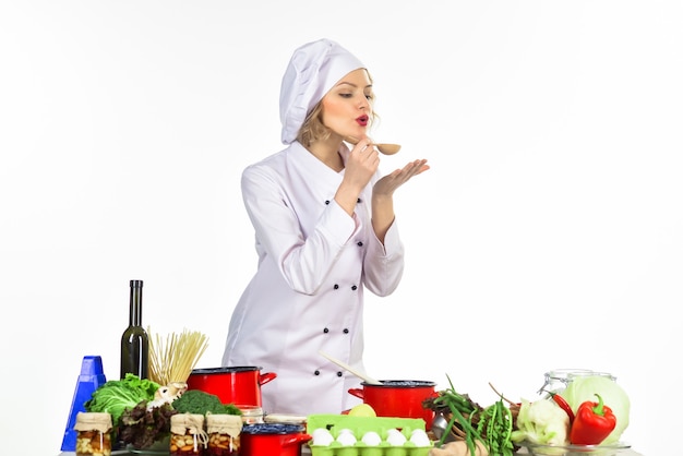 Köchin, die weiße Uniform trägt, hält Holzlöffel mit heißem Essen. Dicke Frau, die Suppe mit Gemüse in der Küche kocht. Professioneller Koch im weißen Anzug, der am Tisch steht. Abendessen kochen - Verkostung.