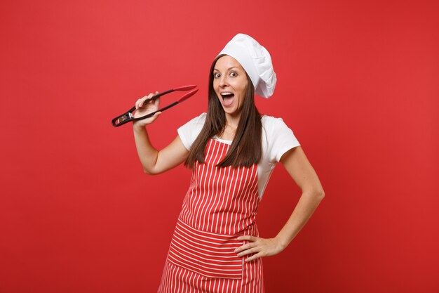 Köchin oder Bäckerin in gestreifter Schürze, weißes T-Shirt Toque Kochmütze isoliert auf rotem Wandhintergrund. Frau hält Küchensalat mit einer Plastikzange zum Grillen. Mock-up-Kopierraumkonzept.