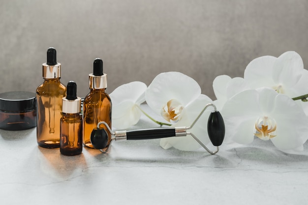 Körper- und Hautpflegeprodukte, Tropfflaschen und Guasha-Massagerolle aus Obsidianstein im Badezimmer mit Orchideenblüten, Markenmodell für Spa-Kosmetikprodukte