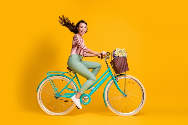 Körpergröße Seitenprofilfoto in voller Länge von fröhlichem Mädchen, das blaues Fahrrad mit Blumenkorb fährt, isoliert auf leuchtend gelbem Hintergrund