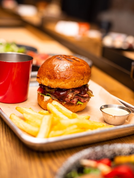 Köstlich lecker gegrillter hausgemachter Burger mit Rindfleisch, Käse, Speck und Sauce auf Holztisch mit Kopierraum. Hände, die Burger mit Pommes Frites und Bier halten Gruppe von Freunden, die Fastfood essen