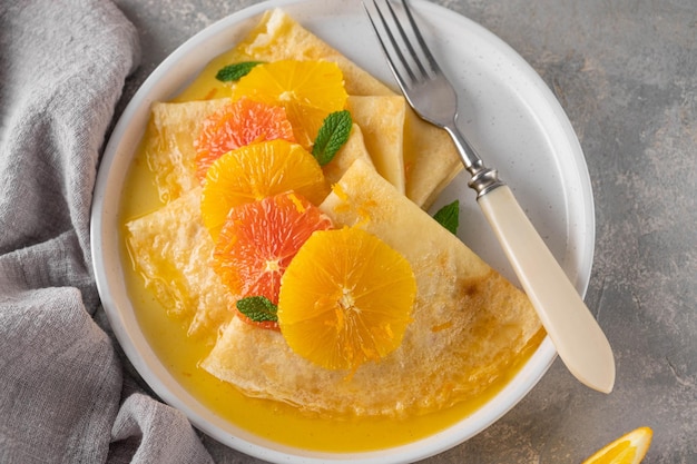 Köstliche Crêpes-Suzette mit Orangensirup und geschnittenen Früchten auf grauem Betonhintergrund