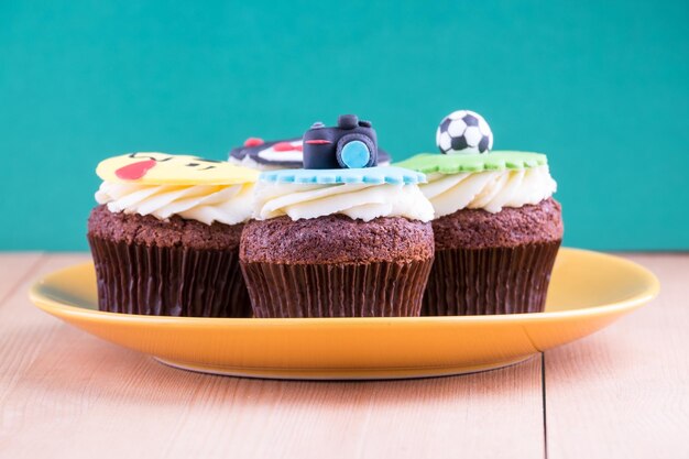 Köstliche Cupcakes mit Ikonen von Ball-Smoking-Smiley und Kamera darauf mit gelbem Teller auf Holzschreibtisch und grünem Hintergrund