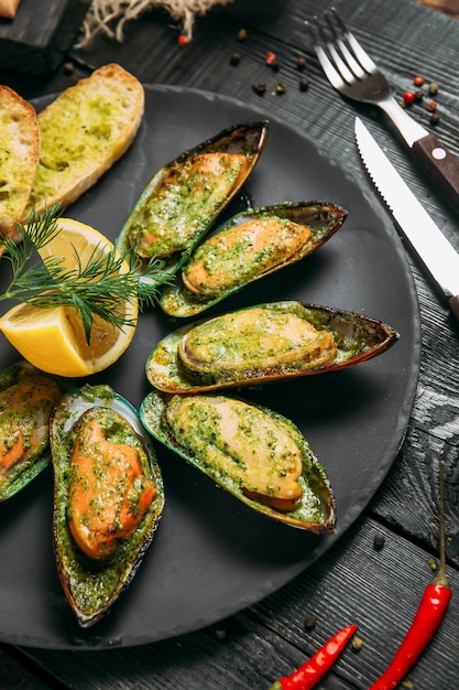 Köstliche gekochte Muscheln mit Soße, Zitronen- und Knoblauchbrot und Butter auf einem schwarzen Teller auf dem dunklen hölzernen Hintergrund