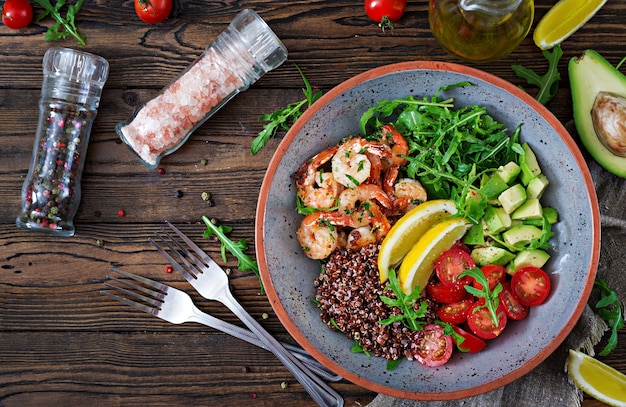 Köstliche gesunde Buddha-Schüssel mit Garnelen, Tomate, Avocado, Quinoa, Zitrone und Arugula auf dem Holztisch. Gesundes Essen. Ansicht von oben. Flach liegen.