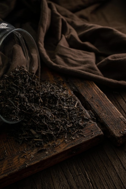 Köstliche getrocknete teeblätter, die aus einem glas auf einem hölzernen vintage-brett auf dunklem hintergrund in der nähe verstreut sind