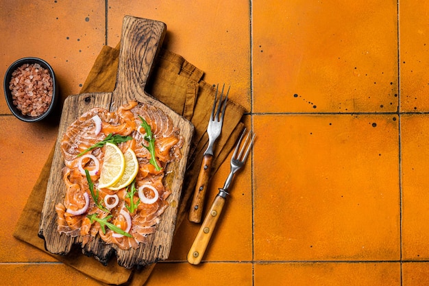 Köstliche Lachs-Carpaccio-Scheiben mit Zwiebeln und Rucola auf einem Holzbrett Orangefarbener Hintergrund Ansicht von oben Kopieren Sie Platz