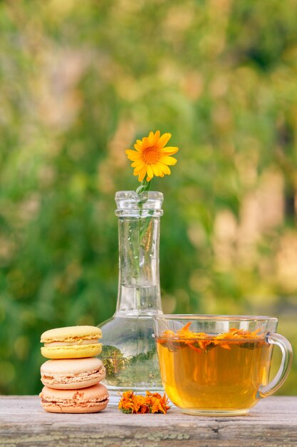 Köstliche Makronenkuchen in verschiedenen Farben, Ringelblume mit einem Stiel in einer Glasflasche und eine Tasse grünen Tee mit trockenen Blumen auf Holzbrettern in unscharfem Naturhintergrund