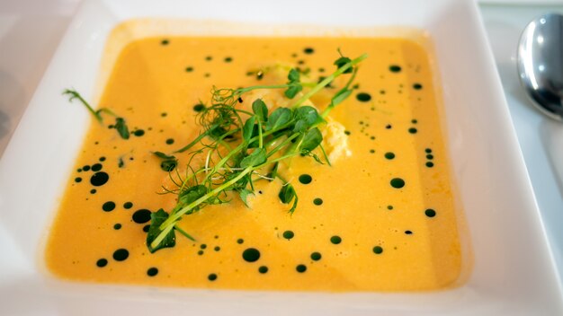 Köstliche orange dicke Kürbispüreesuppe, in weißer quadratischer Schüssel, verziert mit Kräutern auf Tisch.