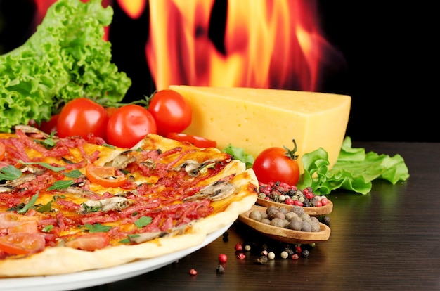 Köstliche Pizza-Salami-Tomaten und Gewürze auf Holztisch auf Flammenhintergrund