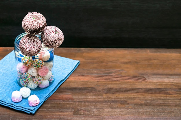 Köstliche Schokoladenkuchen-Pops dekoriert mit weißen Streuseln im Glas mit bunten Bonbons Süßigkeiten auf Serviette Holzhintergrund Gebäckdesserts auf Stick mit Klebeband Leckeres Essen