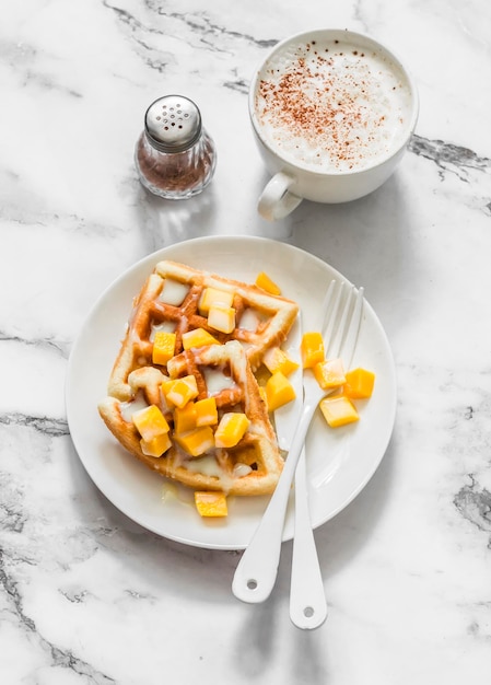 Köstliche süße Frühstückswaffeln mit Mango und Dulce de Leche und Cappuccino auf einer hellen Marmorhintergrund-Draufsicht