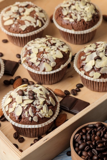 Köstliche, süße Schokoladenmuffins mit Mandelblüten in einem Holztablett neben Kaffeekörnern auf einem blauen Holztisch.