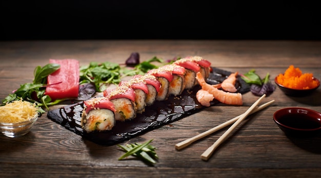 Köstliche Sushi-Rollen mit Reis und Fisch auf schwarzem Schiefer, serviert auf dem Holztisch mit Essstäbchen, Schüssel mit Sojasauce, Kaviar und Sprossen