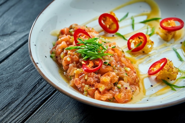 Köstliche Vorspeise - Lachstatar mit Avocado und scharfem Chili auf einem weißen Teller auf schwarzer Holzoberfläche.