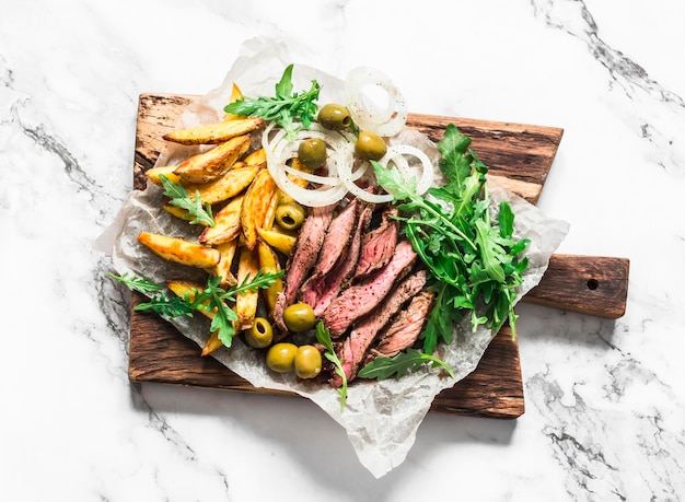 Köstliche Vorspeisen an Bord von mittelhartem Rindersteak, Pommes Frites, grüner Salat, Oliven, eingelegte Zwiebeln auf hellem Hintergrund, Draufsicht