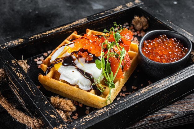 Köstliche Waffel mit geräuchertem Lachsfilet, pochiertem Ei und rotem Kaviar. Schwarzer Hintergrund. Ansicht von oben.