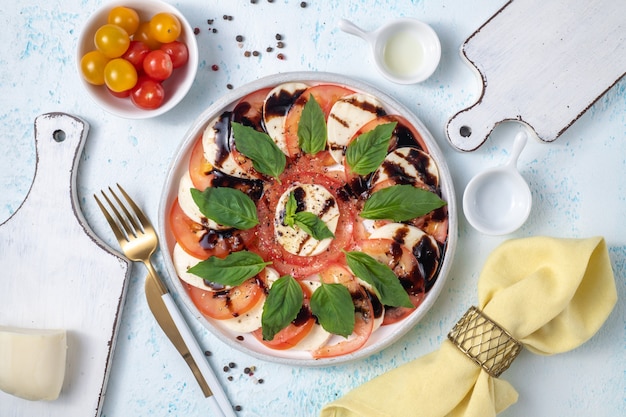 Köstlicher Caprese-Salat mit reifen Tomaten und Mozzarella-Käse, frischen Basilikumblättern, Olivenöl und Balsamico-Sauce. Italienisches Essen. Flache Draufsicht auf blauem Hintergrund