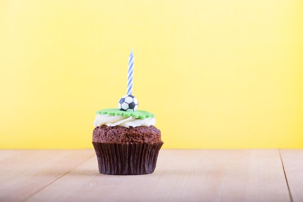 Köstlicher Cupcake mit Kugelsymbol darauf und Kerze auf Holzschreibtisch und gelbem Hintergrund