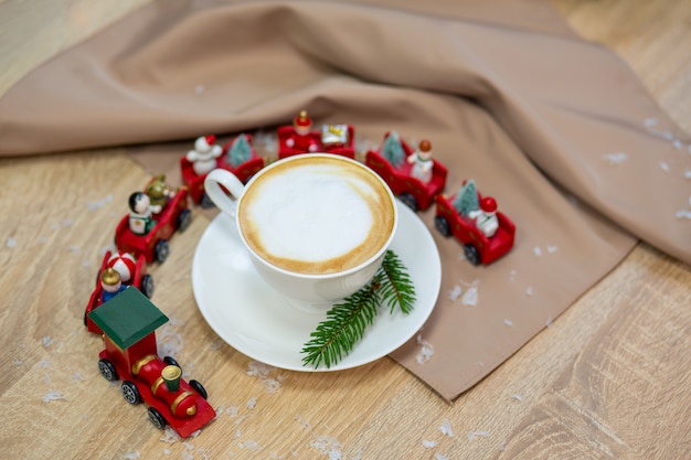 Köstlicher frischer festlicher Morgencappuccinokaffee in einer keramischen weißen Schale auf dem Holztisch mit dekorativem Weihnachtszug, roten Ornamentals, Leuchtkäfern und Fichtenzweigen.