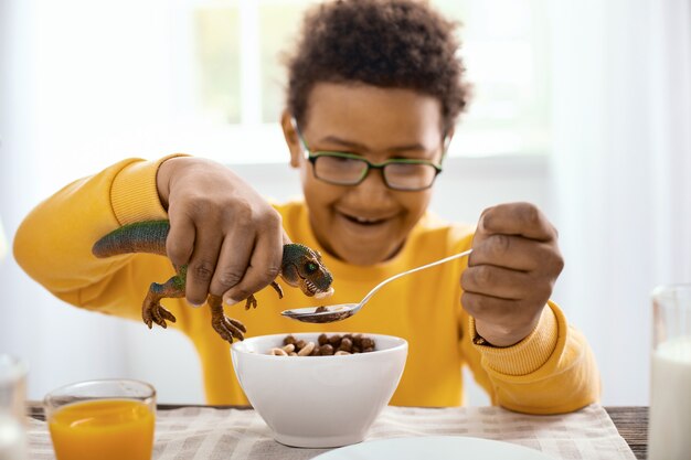 Köstlicher Snack. Angenehmer jugendlicher Junge, der Müsli zum Frühstück isst und sie seinem Spielzeugdinosaurier füttert, während er lächelt