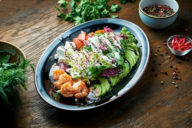 Köstlicher und diätetischer Salat mit gegrillten Garnelen, Feta-Käse, Tomaten und Avocado, serviert in einem schwarzen Teller auf einem Holztisch. Restaurant Essen