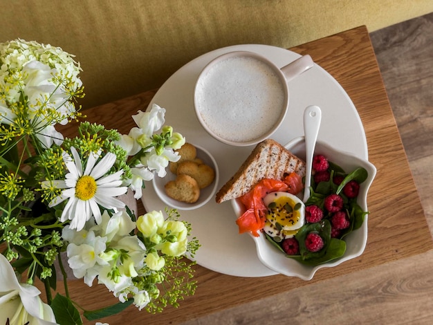 Köstliches Frühstück auf einer Holzbank im Wohnzimmer Cappuccino Lachs Toast gekochtes Ei Himbeer Spinat Salat