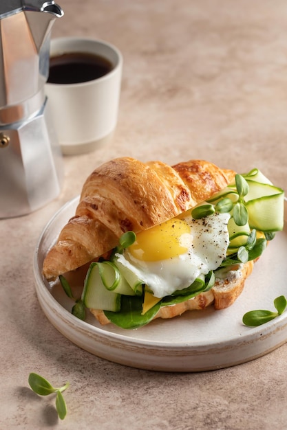 Foto köstliches frühstück mit croissant-sandwich mit spiegelei-käse-gurke und mikrogemüse und kaffee auf beigem hintergrund. gesundes, köstliches frühstück mit textfläche