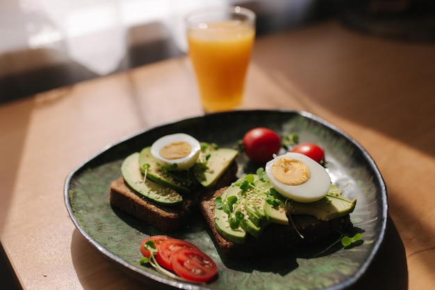 Köstliches Frühstück zu Hause Sandwich mit frisch geschnittener Avocado über geröstetem Roggenbrot mit Kirschtomaten und gekochtem Ei auf grüner Platte Frischer Orangensaft