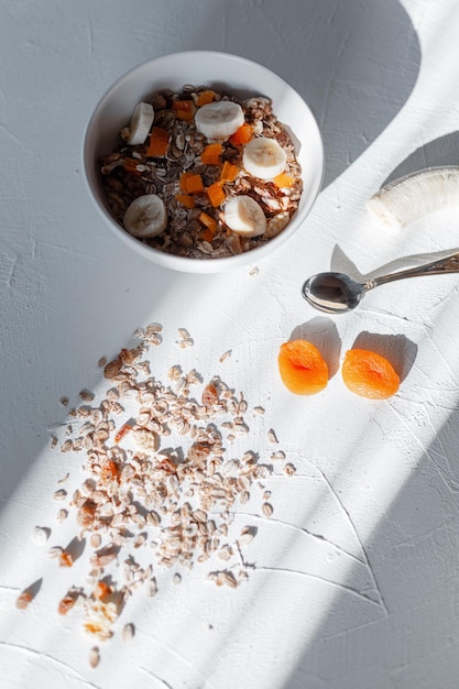 Köstliches, gesundes Frühstücksmüsli mit Haferflocken, getrockneten Aprikosen und Bananen auf einem weißen Tisch bei Sonnenaufgang am Morgen. Vitamine und natürliche Rohkost. Veganes Konzept