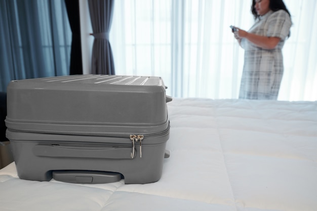 Koffer auf dem Bett im Hotelzimmer und junge Frau, die den Transfer über die mobile Anwendung bestellt,
