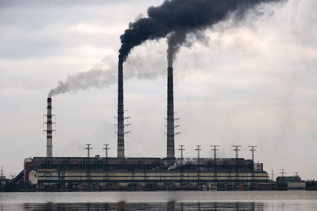 Kohlekraftwerk hohe Rohre mit schwarzem Rauch, der sich nach oben bewegt, verschmutzende Atmosphäre mit Reflexionen davon im Seewasser.