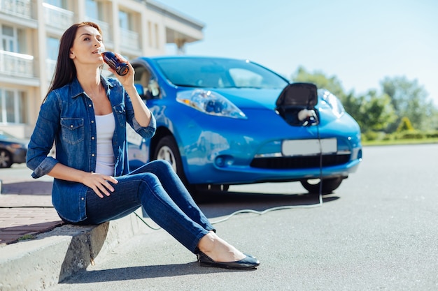 Kohlensäurehaltiges Getränk. Positive brünette nette Frau, die eine Flasche hält und Soda trinkt, während sie vor ihrem Auto sitzt