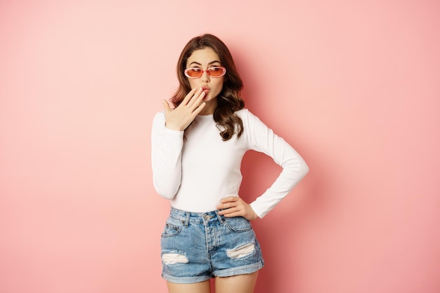 Kokettes und stilvolles brünettes Mädchen, lachend und lächelnd, mit Sonnenbrille, weißer Bluse und Jeans, rosa Hintergrund