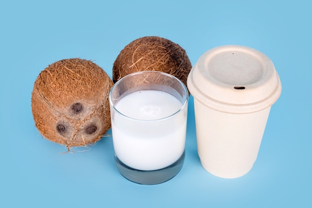 Kokosnüsse mit einem Glas Kokosmilch und Papierkaffeetasse auf blauem Hintergrund. Gesundes veganes Essen, laktosefreie Ernährung, Milchalternative, Coffee to go, veganer Latte oder Kokos-Cappuccino.