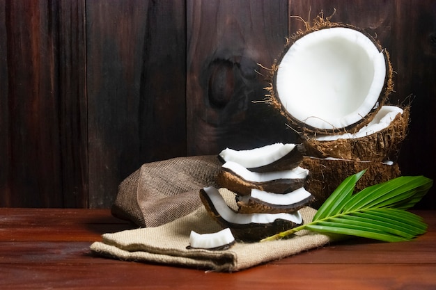 Kokosnusshälfte und Kokosnussstücke und -blatt auf Holztisch und schwarzem hölzernem Hintergrund