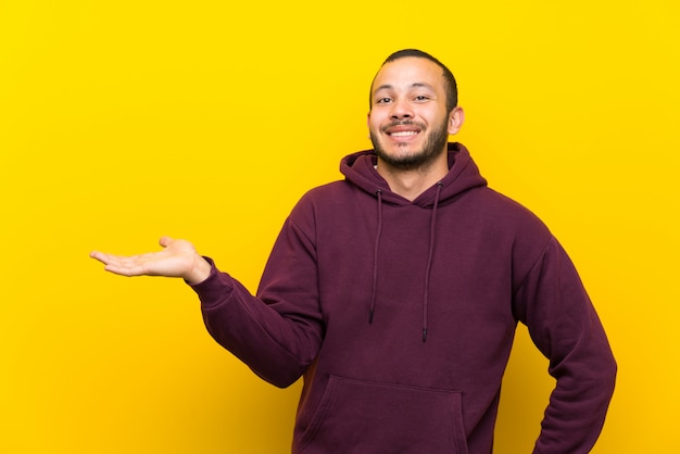 Kolumbianischer Mann mit Sweatshirt über der gelben Wand, die copyspace eingebildet auf der Palme hält